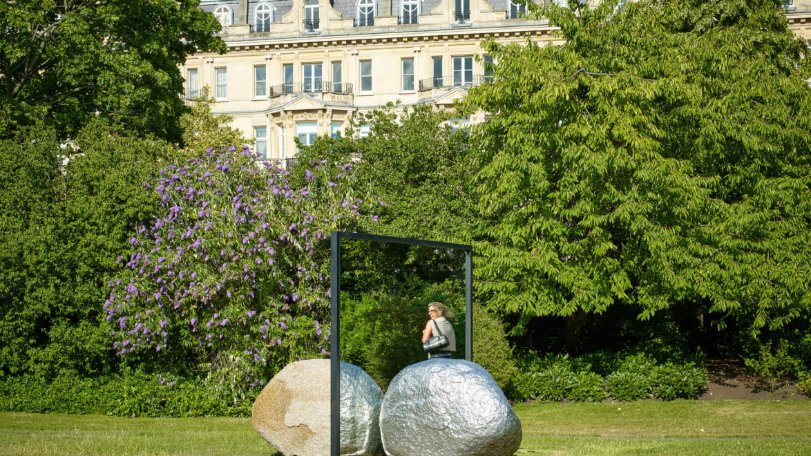 Outdoor sculpture of a person with a mirror among trees in a park, with large building in background on a sunny day.