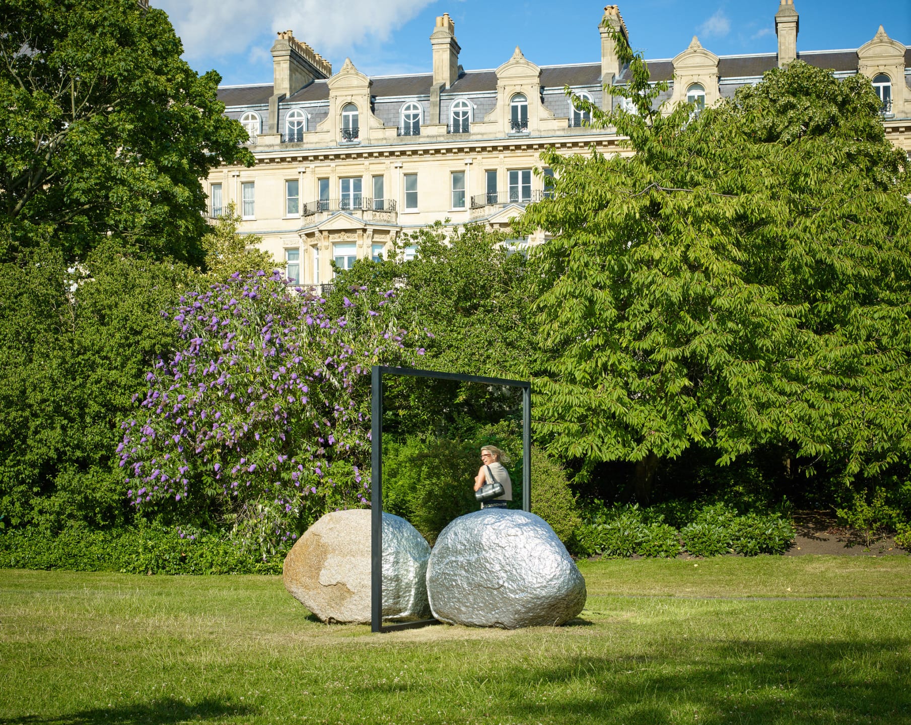Outdoor sculpture of a person with a mirror among trees in a park, with large building in background on a sunny day.