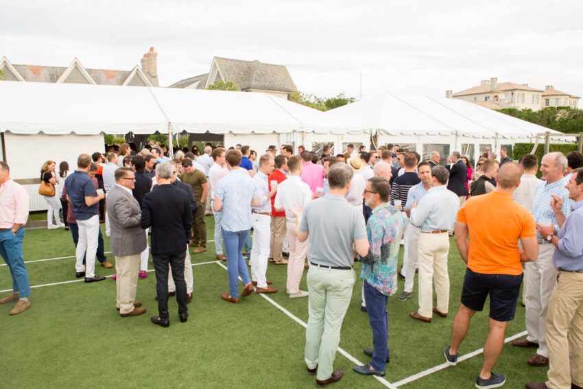People socializing at an outdoor event on a grassy area with white tents in the background.