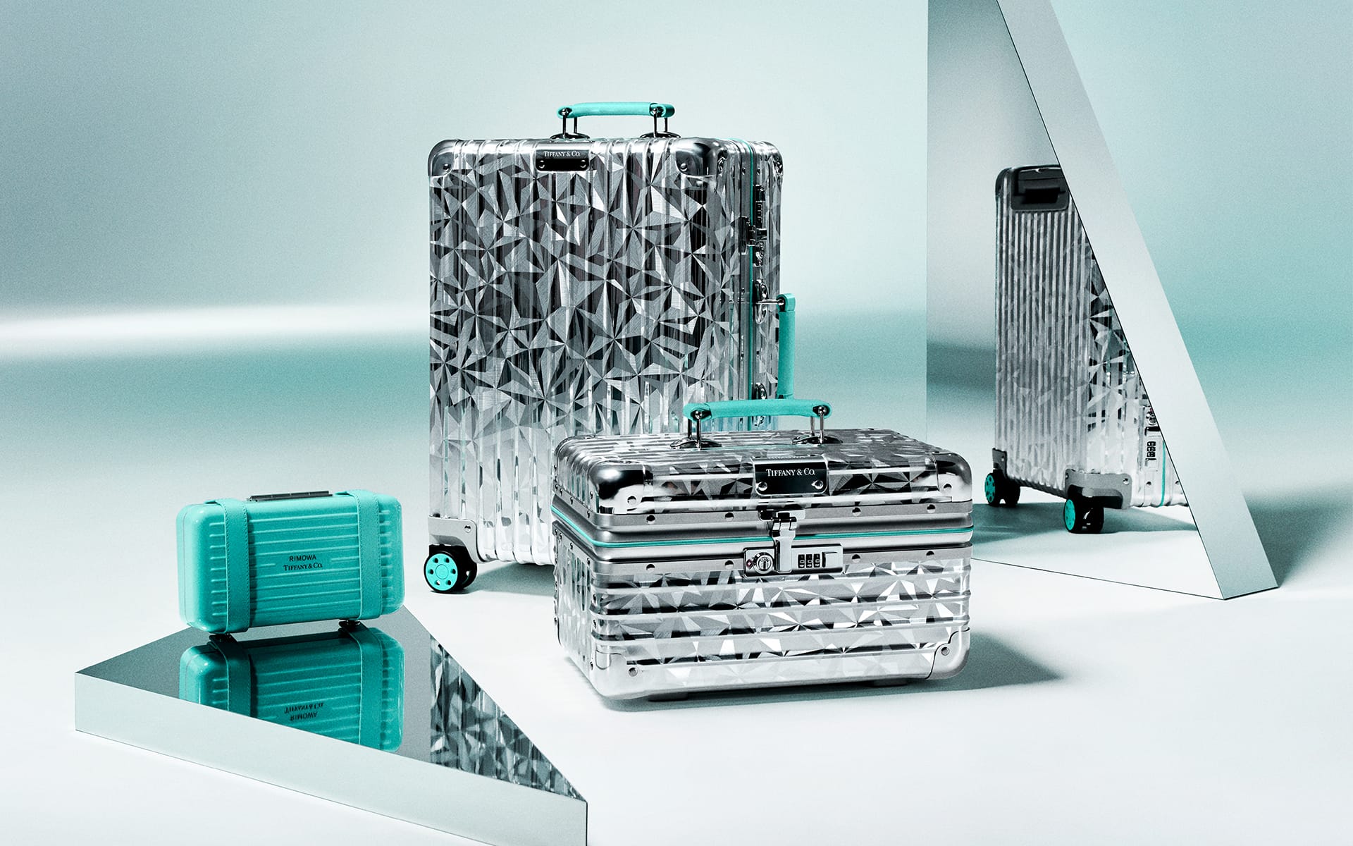 Shiny geometric-patterned silver luggage set with turquoise accents displayed on mirrored surfaces in a minimalist setting.
