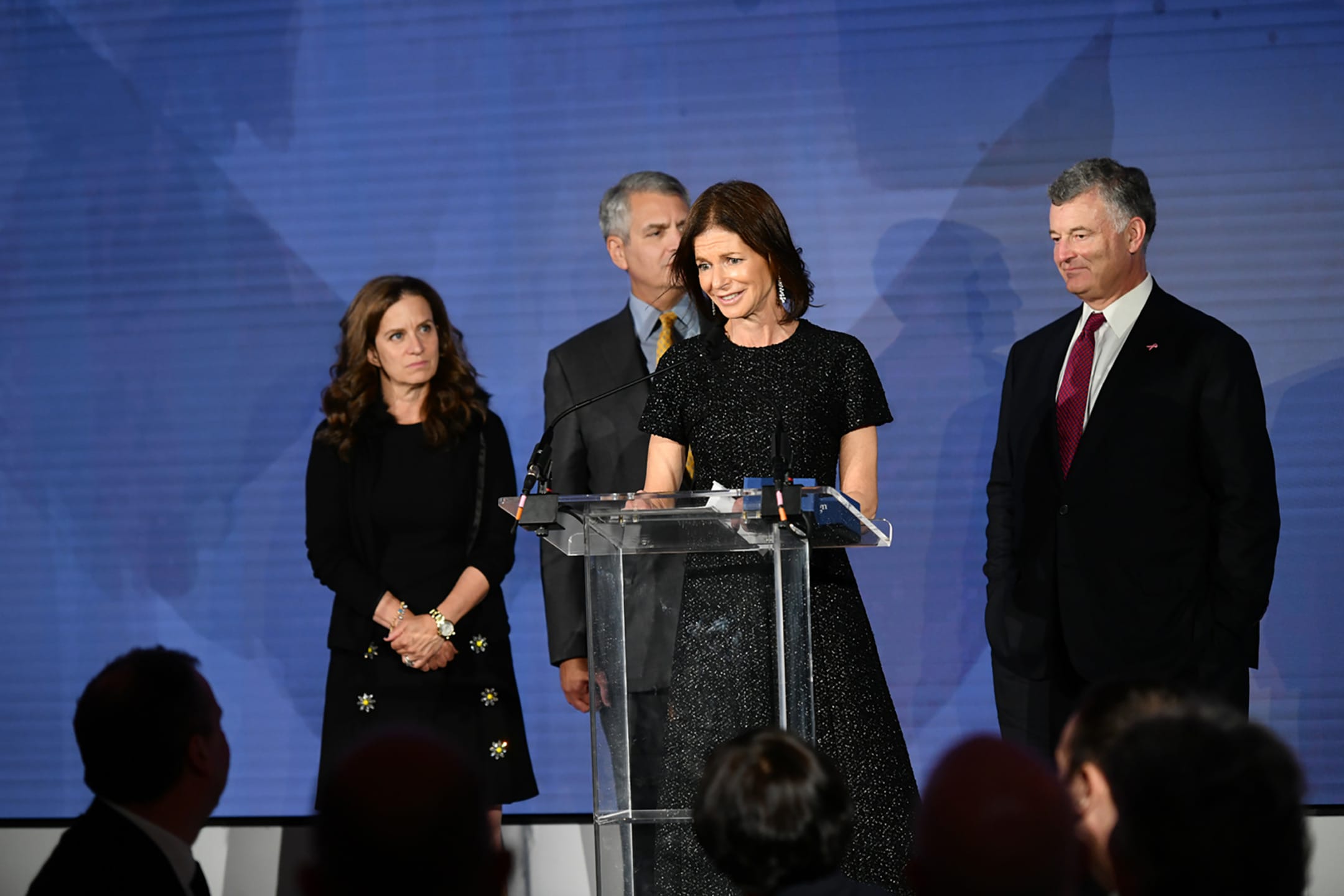 Four people on stage, one woman speaking at a clear podium, others standing and listening, large screen backdrop.