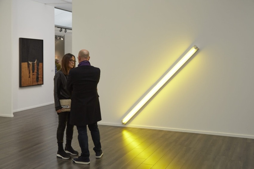 Two people stand in an art gallery discussing a yellow neon light installation on a white wall.