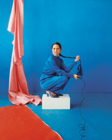 Woman in blue dress holding long braid, sitting on a white block, surrounded by blue, red, and pink fabric.