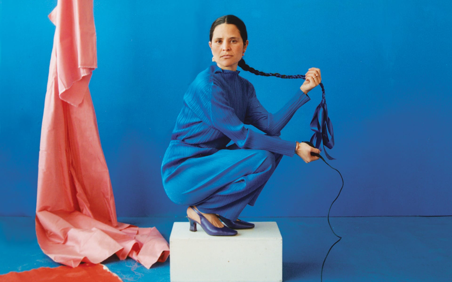 Person in a blue outfit with blue shoes, squatting and holding a braided rope, with pink fabric draped in the background.