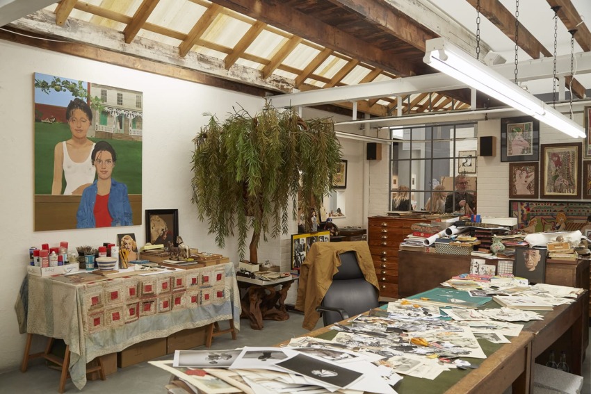 Artist's studio with paintings, an indoor tree, scattered photos on a table, art supplies, and overhead lighting.