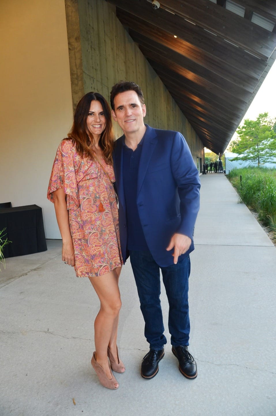A man and a woman posing and smiling in front of a modern building with a slanted roof.