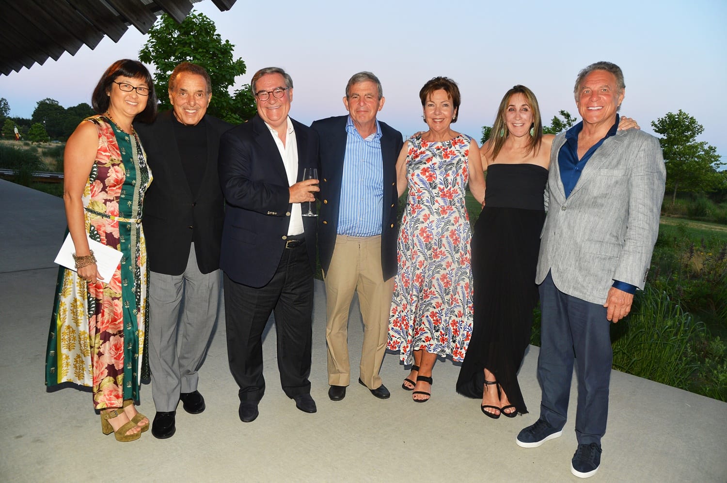 Group of seven people posing together outdoors on a patio, dressed in formal attire, with trees and sky in the background.