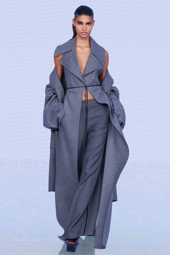 Model wearing a long gray coat and pants on a fashion runway.