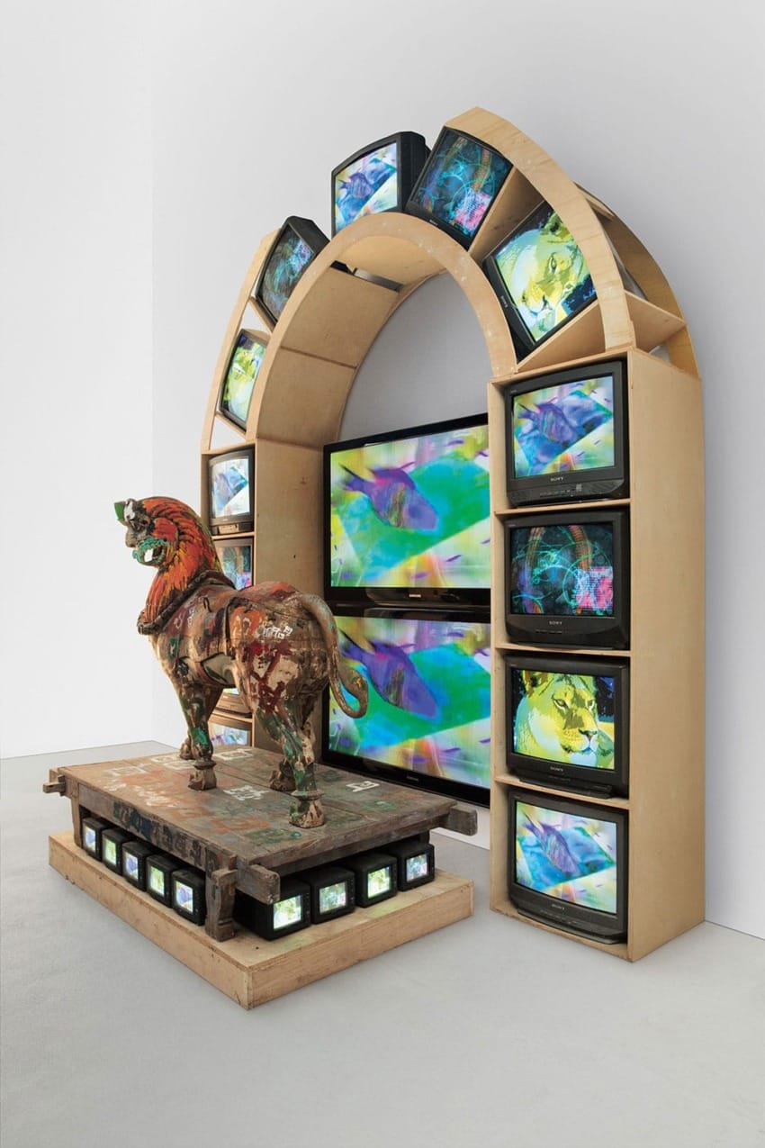 Art installation with a wooden arch of TV screens displaying colorful visuals and a painted lion statue on a platform.