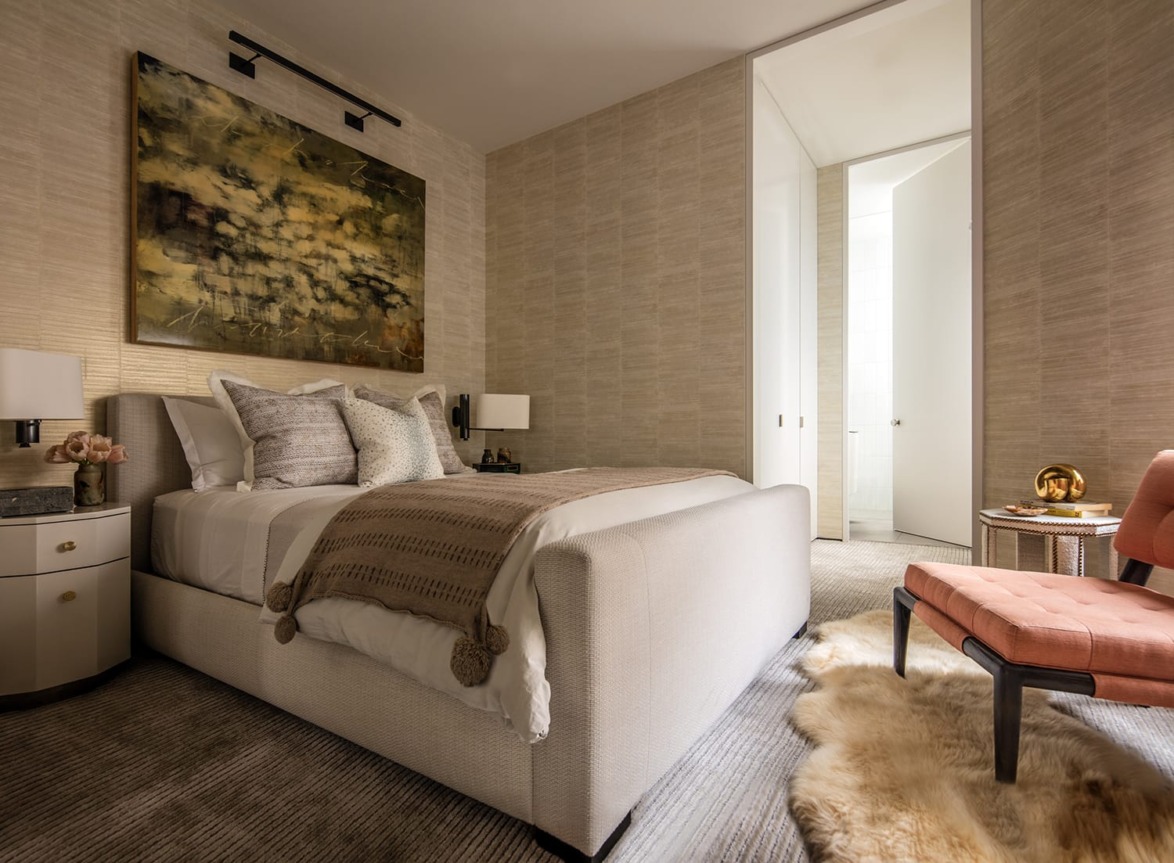 Elegant bedroom with cozy bed, textured wallpaper, abstract art, soft lighting, and an inviting chair with a fur rug.