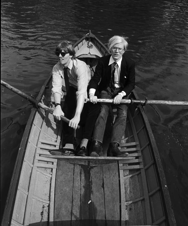 Two men in suits rowing a wooden boat on a lake.