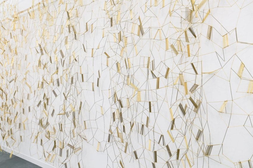 Intricate geometric wire art installation on white wall with gold and silver intersecting lines creating abstract patterns.