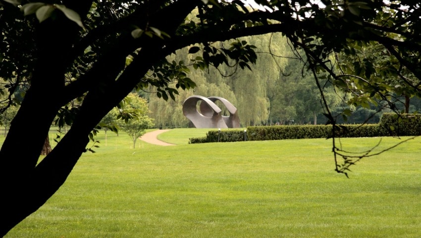 Curved sculpture on a manicured lawn framed by tree branches in a park setting.