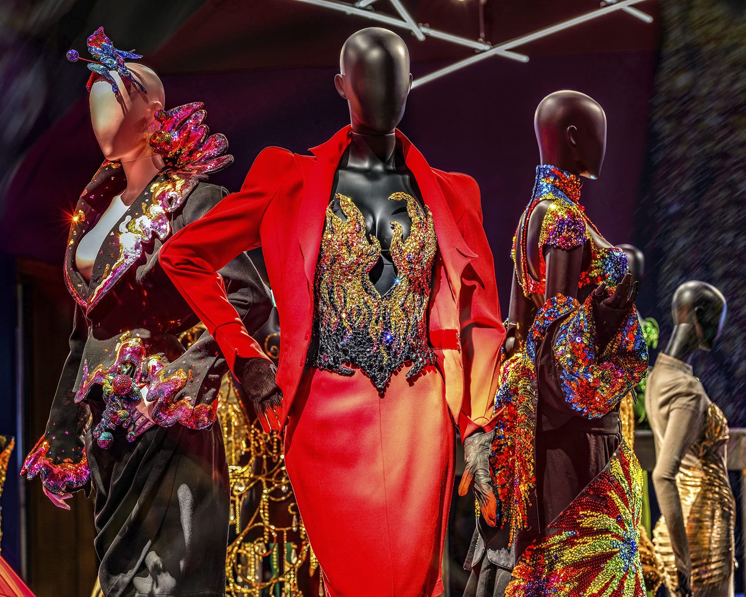 Mannequins showcasing vibrant, sequined gowns and jackets under bright lighting in a fashion display.