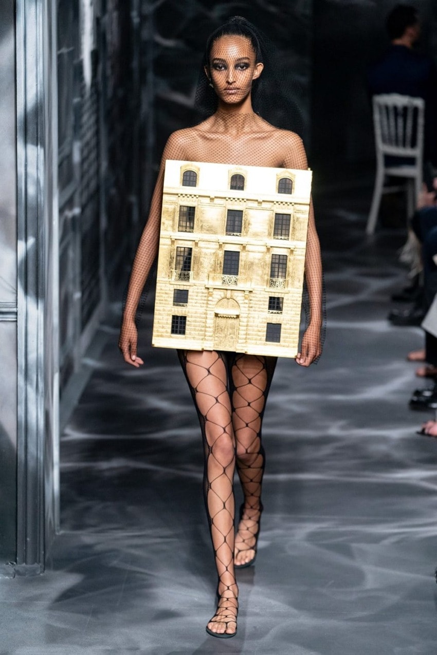 Model on runway wearing unique building-themed outfit and fishnet stockings.