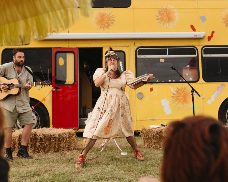 Person in vintage dress singing on stage with guitar player by yellow bus in outdoor setting.
