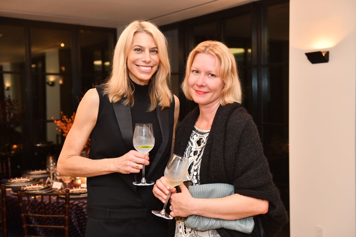 Two people standing indoors, both holding wine glasses and smiling, with a dinner table and large windows in the background.