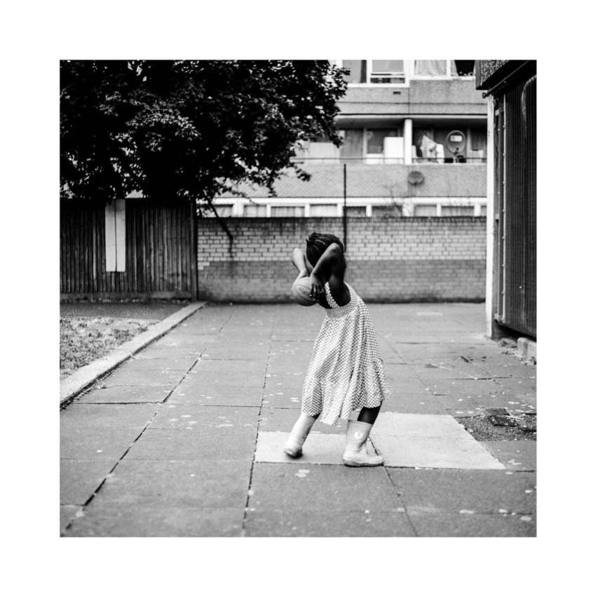 Child dancing in polka dot dress outdoors on a paved area, surrounded by buildings and trees in black and white.