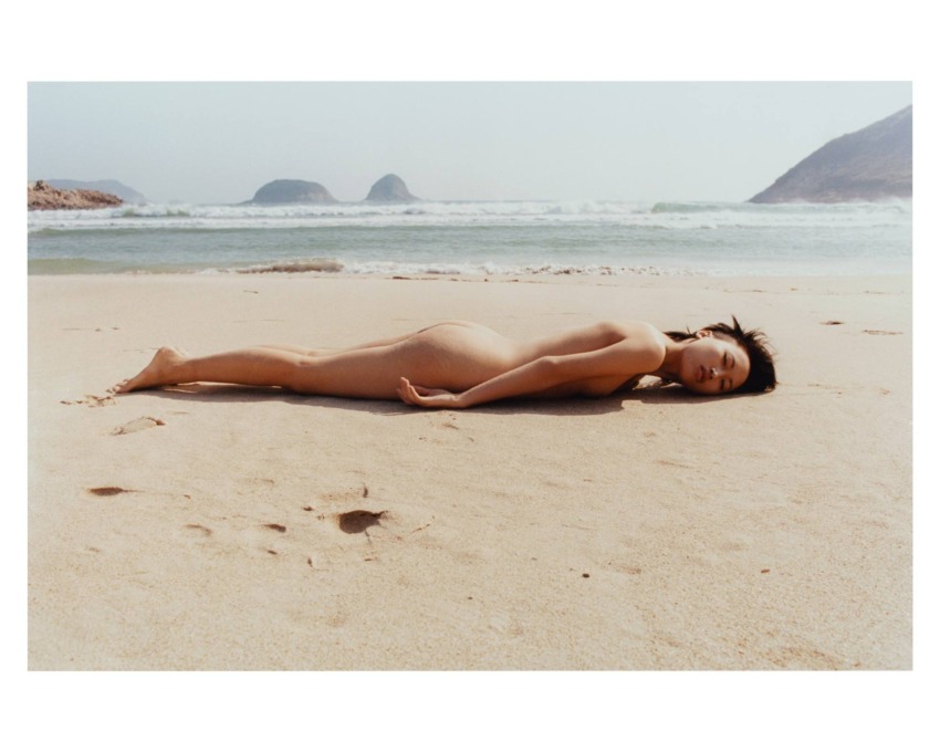 Person lying on sandy beach with ocean and distant hills visible in the background on a cloudy day.