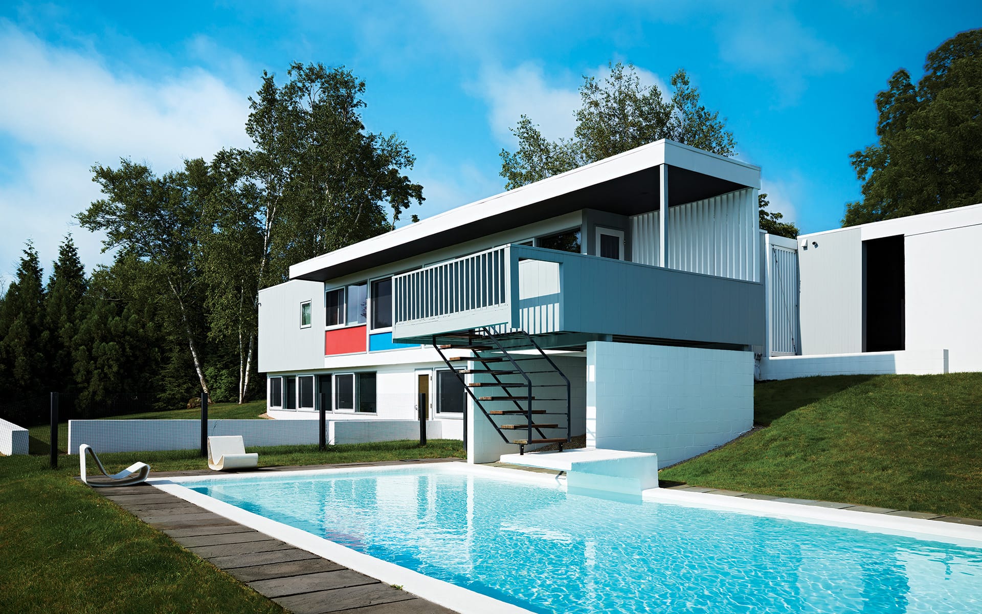 Modern house with colorful accents, elevated terrace, and a rectangular pool surrounded by trees and green lawn.