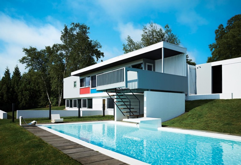 Modern house with flat roof, large windows, colorful accents, surrounded by trees, and a swimming pool in the foreground.