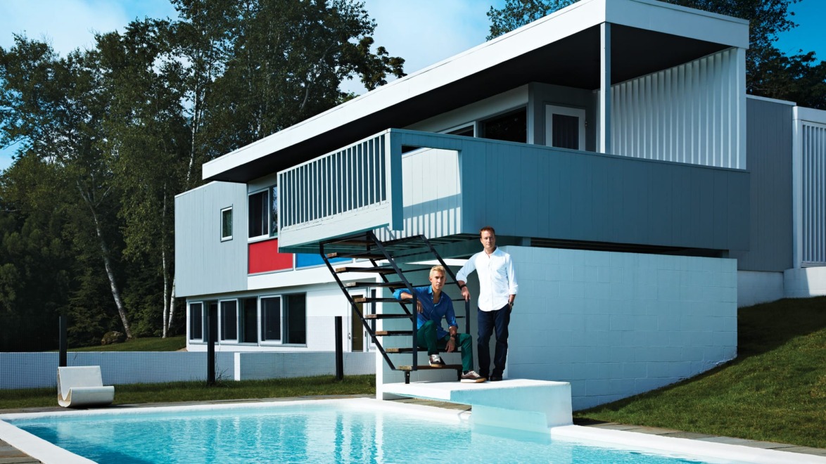 Two people standing on steps outside a modern house with a pool in front, surrounded by trees.