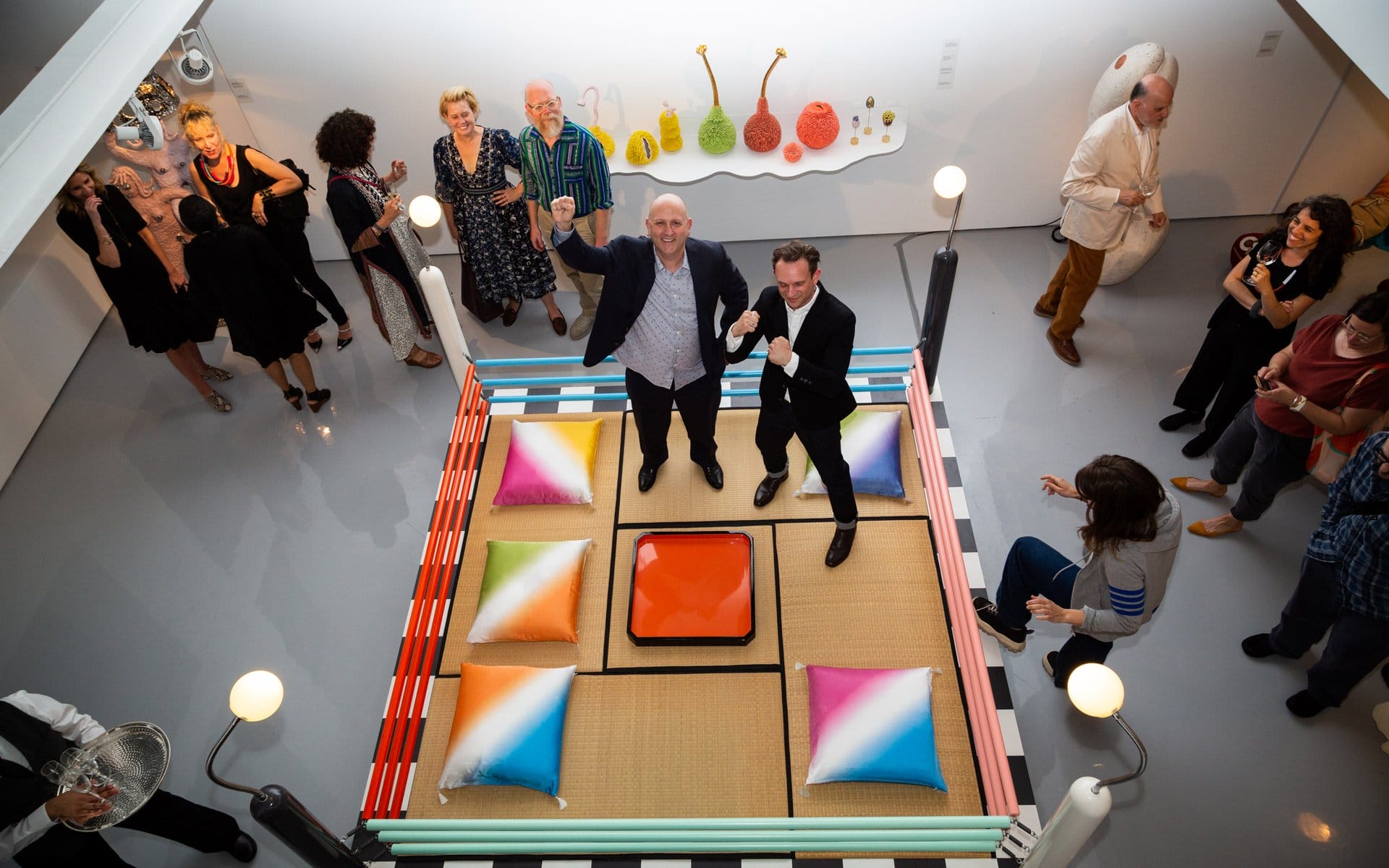 A group of people socializing around a colorful, square, pillow-filled platform at an art gallery event.
