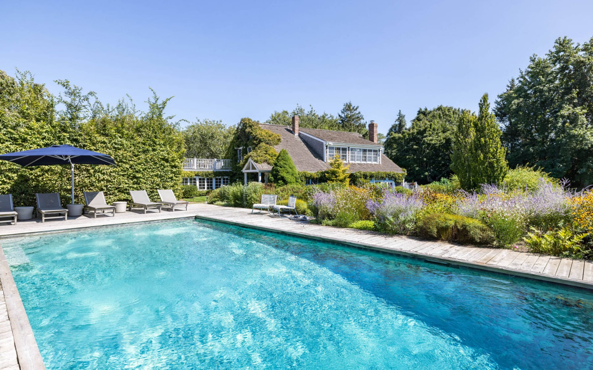 Luxurious outdoor pool with sun loungers and umbrella, surrounded by greenery and a charming country house in the background.
