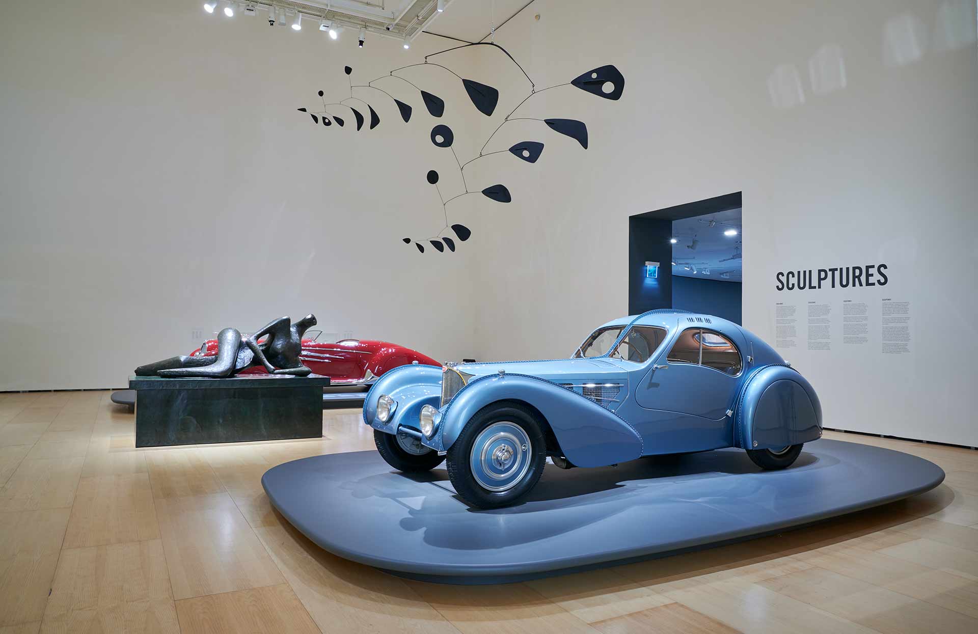 Car exhibit with blue vintage car and abstract art sculpture in a modern gallery setting