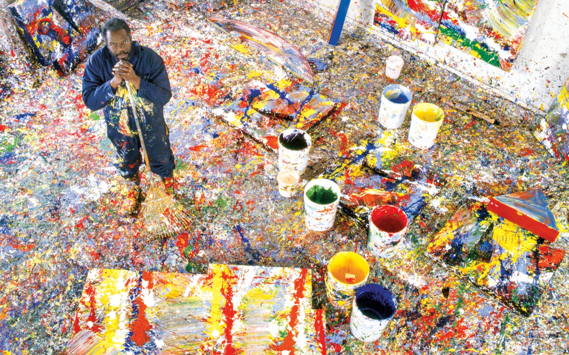 Artist standing in a studio surrounded by colorful paint splatters and large canvases.