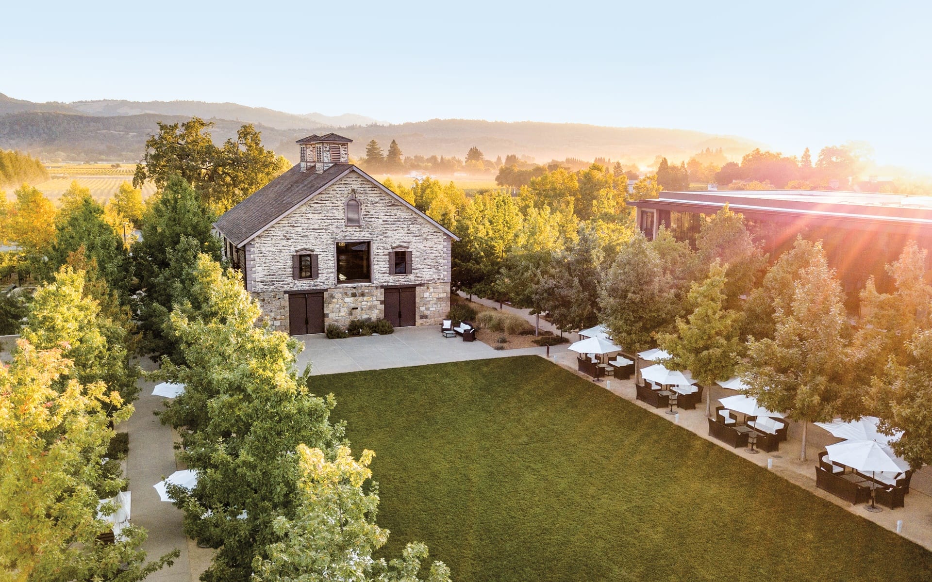 Rustic stone barn surrounded by lush trees and green lawn, with scenic mountains and sunset in the background.
