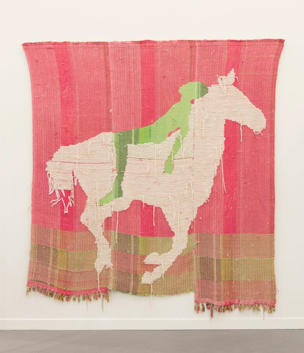 Colorful woven tapestry featuring a white horse and green rider against a pink background, displayed on a gallery wall.