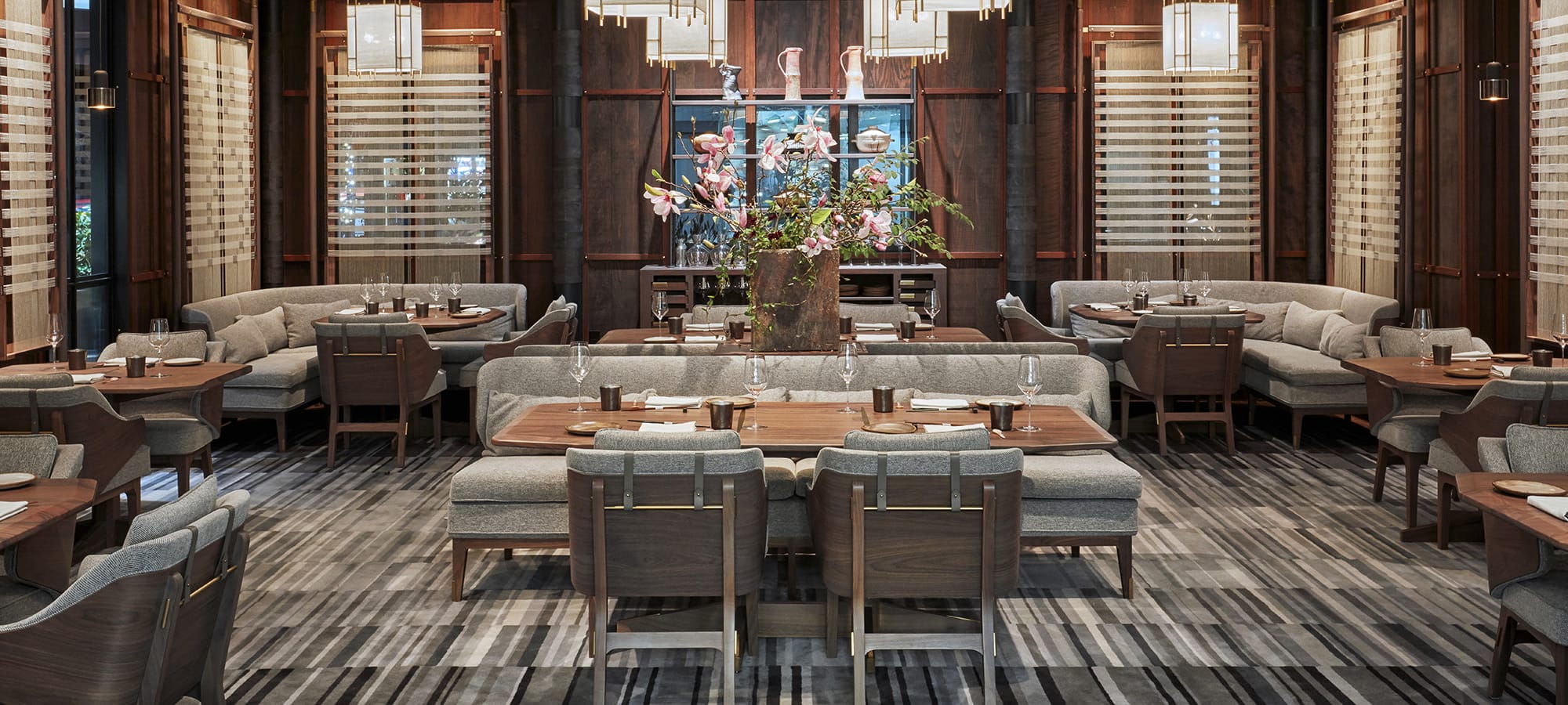 Elegant modern restaurant interior with wooden tables, gray upholstered chairs, and a central floral arrangement.