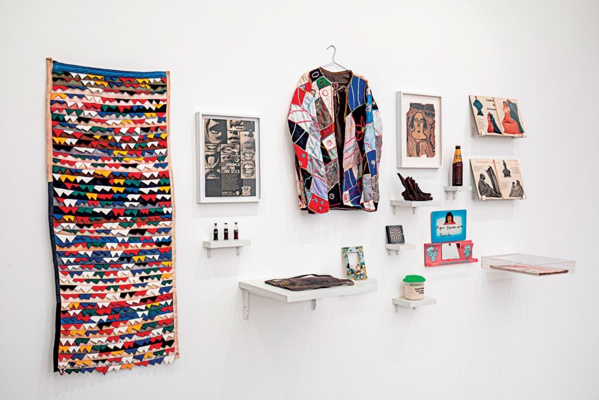 Colorful art exhibition with a woven textile, patchwork jacket, framed artworks, and small figurines displayed on a white wall.