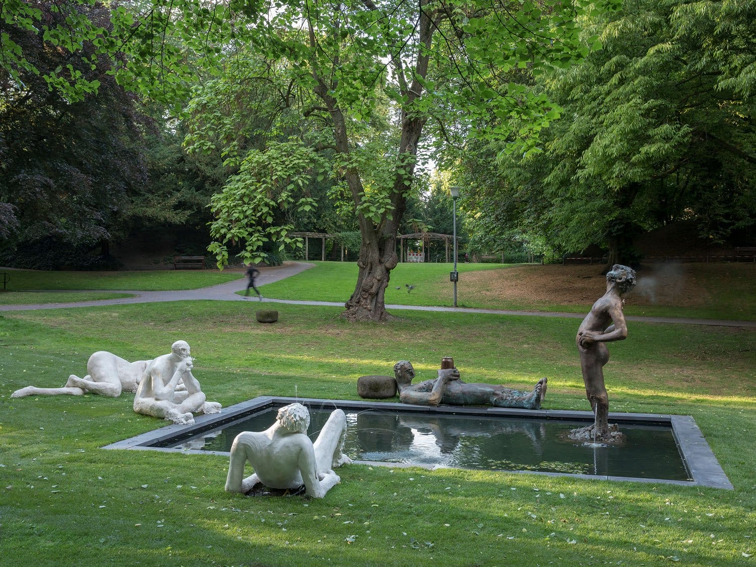 Sculptures in a park surrounding a small pond, with lush green trees and a walking path in the background.