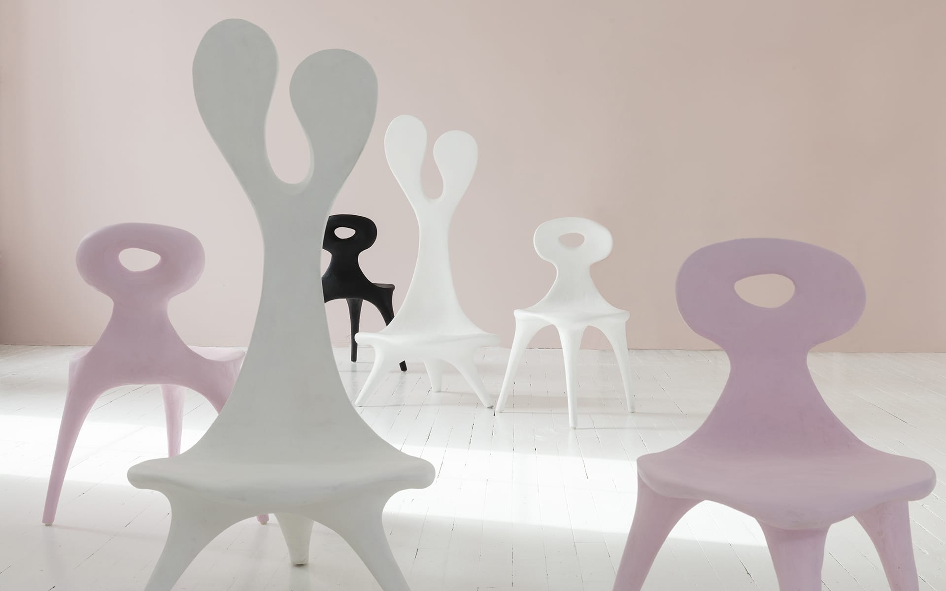 Five modern, uniquely shaped chairs in pastel shades of pink, white, and black on a white floor and pink wall background.