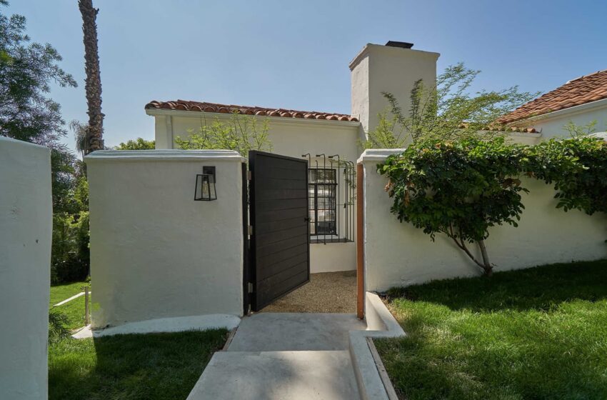 Wooden gate entrance to a white stucco house with greenery and a tiled roof on a sunny day.