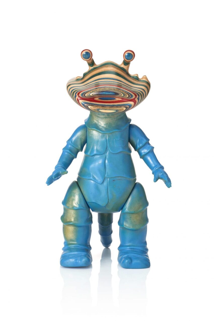 Colorful abstract toy figure with a spiral-patterned head and blue body, standing upright on a reflective white surface.
