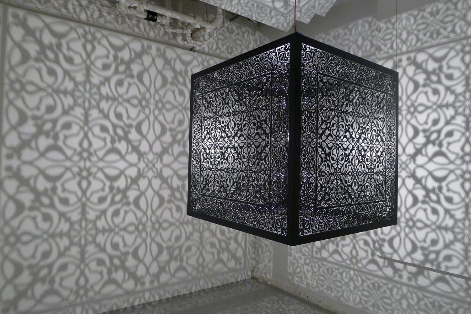 Intricate black cube sculpture casting complex shadows on a white wall in an art gallery setting.