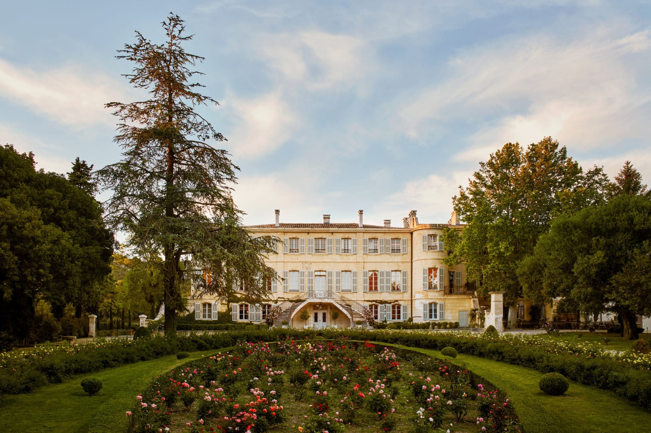 Historic mansion with grand garden and vibrant flowers under a blue sky with clouds, surrounded by trees.