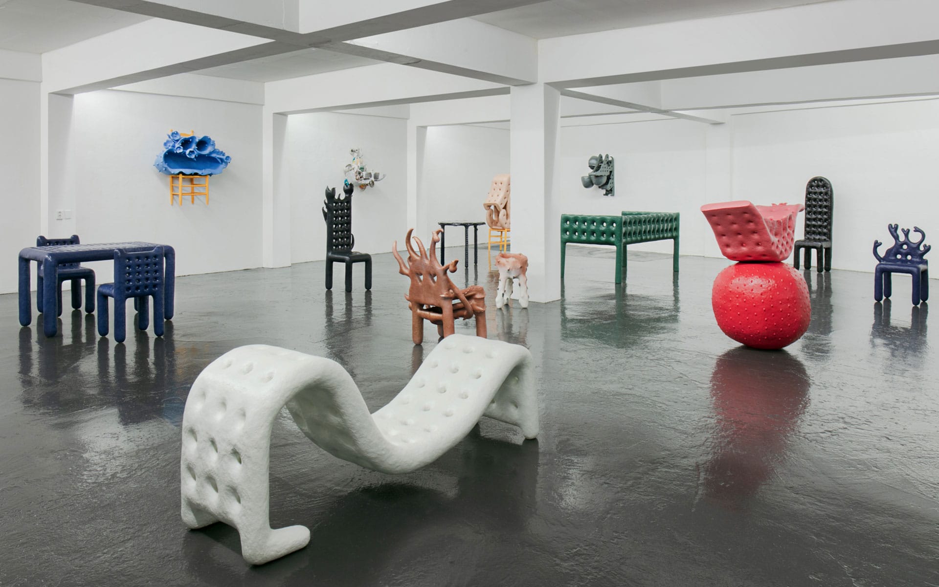 Modern art exhibition featuring abstract, colorful sculptural furniture in a white gallery space with a reflective floor.