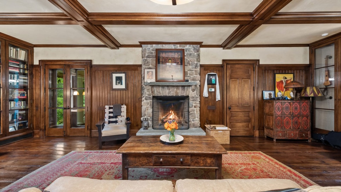 Cozy living room with a stone fireplace, wooden beams, a red rug, and bookshelves filled with books.