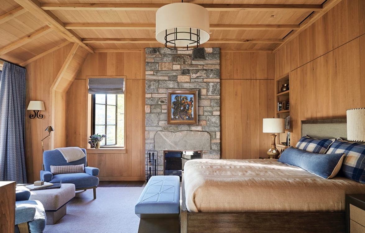 Cozy bedroom with stone fireplace, wooden walls, and large bed. Blue chair and ottoman by the window.