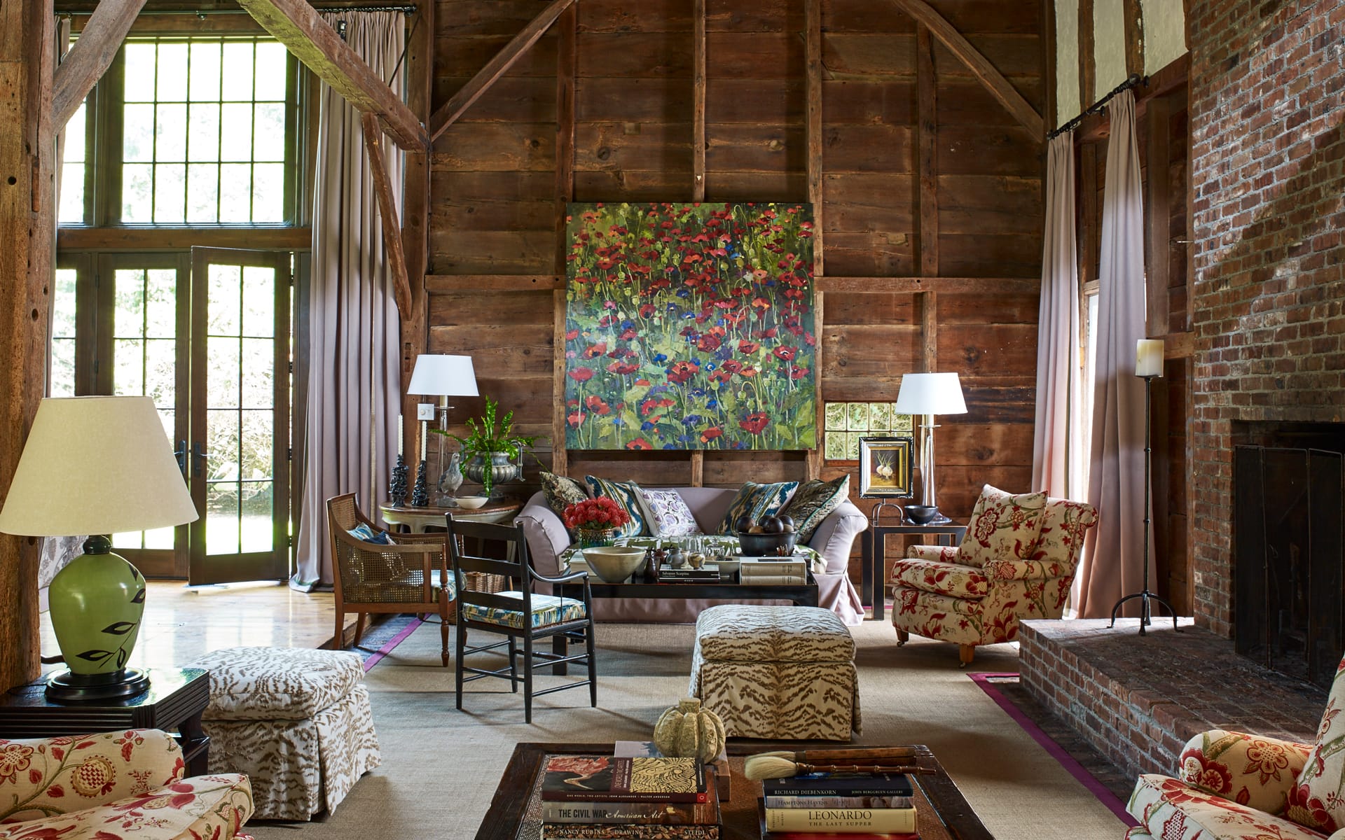 Cozy living room with floral artwork, patterned furniture, wooden walls, large windows, and a brick fireplace.