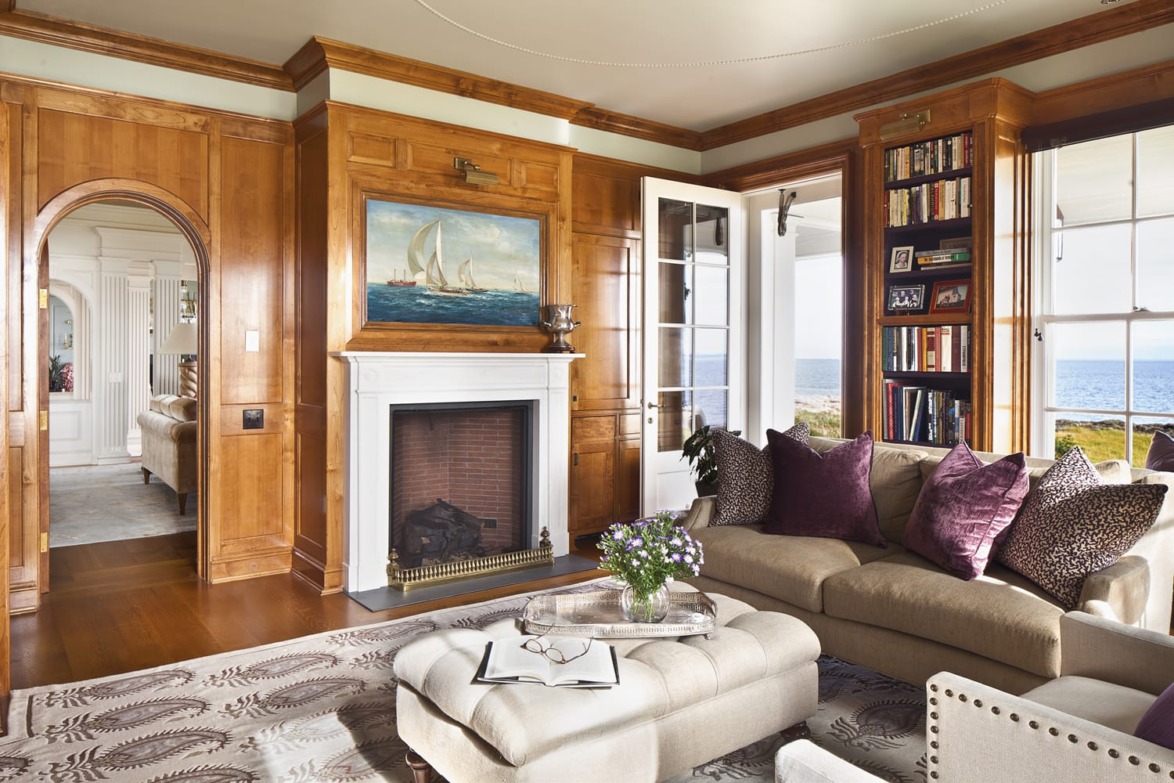 Cozy living room with a fireplace, wooden wall panels, plush furniture, bookshelves, and ocean view through large windows.