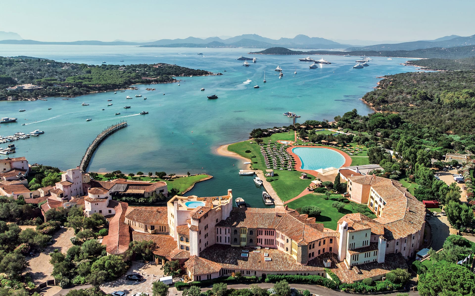 Aerial view of coastal resort with lush greenery, luxury buildings, pools, and numerous boats on a vibrant blue sea.