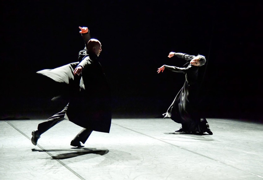 Two dancers in flowing costumes performing expressive movements on a dimly lit stage.