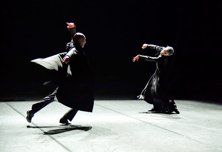 Two dancers in flowing costumes performing expressive movements on a dimly lit stage.