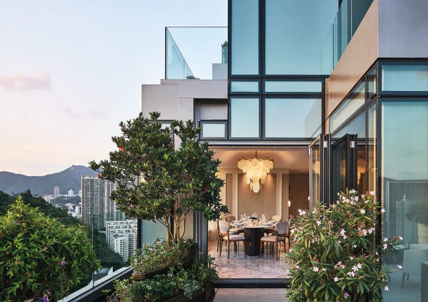 Modern glass-walled dining room with city skyline view, greenery, and outdoor plants at sunset.