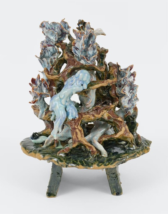 Ceramic sculpture of a blue figure surrounded by abstract, leaf-like structures on a round base with legs.